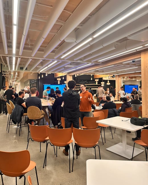 A candid photograph captures a large gathering of people in a modern, open-plan indoor space characterized by high wooden ceilings, linear lighting, and numerous orange molded chairs.