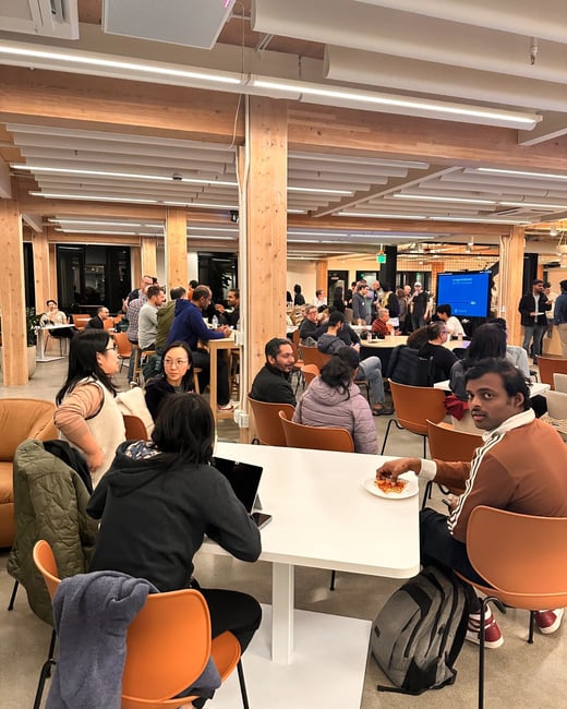 A candid photograph captures a large group of people socializing and eating in a modern, brightly lit communal office space featuring wooden structural elements and white ceilings. Several individuals are seated at white tables with orange chairs, enjoying what appears to be a casual dinner or event.