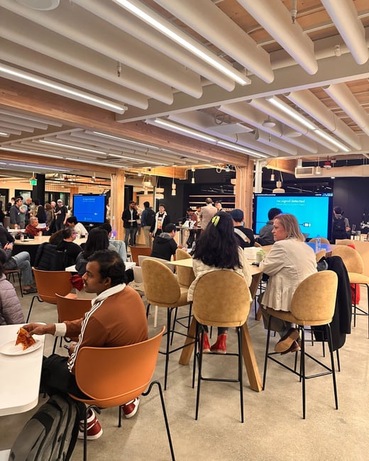 A candid photograph captures a large group of people socializing and eating in a modern, brightly lit indoor space with high ceilings and wooden accents. Attendees are gathered around high tables, and two large display screens are visible in the background, one of which shows a "no signal detected" message.