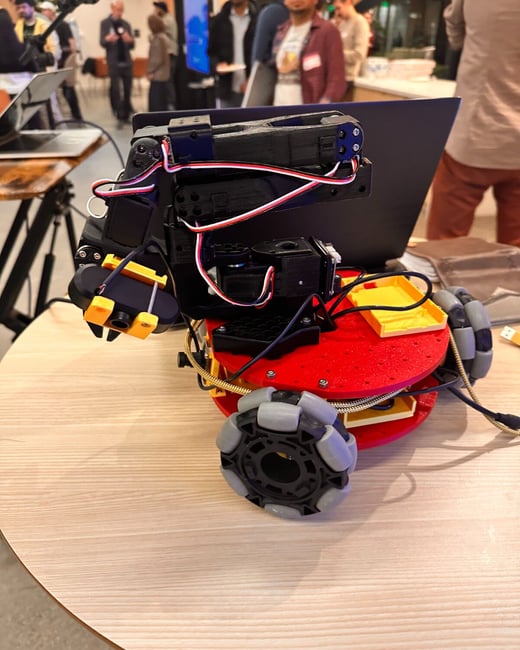 A close-up photograph focuses on a small, custom-built mobile robot prototype with a red base, black components, and grey mecanum wheels, sitting on a light wood table. The background is blurred, showing several people standing and mingling at an indoor event.
