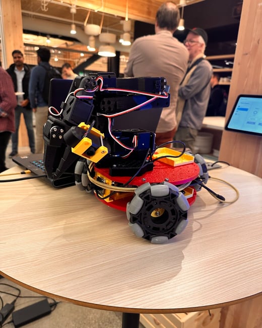 A close-up photograph of a small, complex robotic prototype, featuring red, black, and yellow components and specialized wheels, displayed on a light wooden table during an indoor technology gathering. Several people are visible socializing in the blurred background.