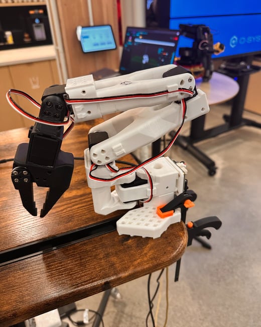 A close-up photograph shows a small, white and black robotic arm prototype secured to a wooden desk, with multiple computer screens and equipment visible in the blurred background of an indoor lab or office.