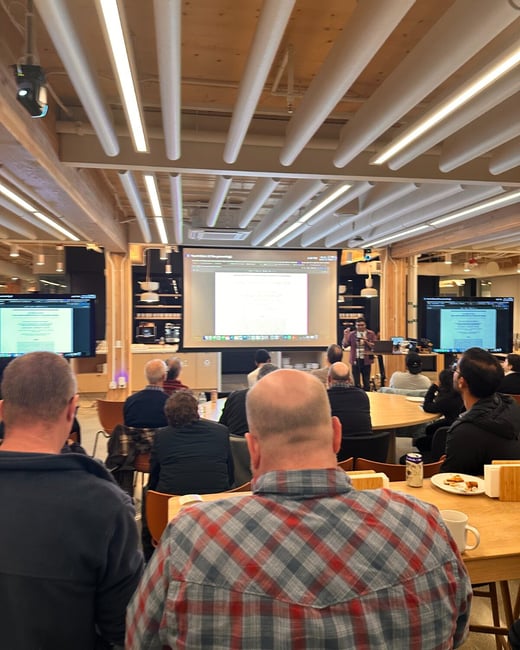 A photograph taken from the audience's perspective shows a group of people attending an indoor presentation, where a speaker stands in front of a projection screen and flanking monitors in a modern office space.
