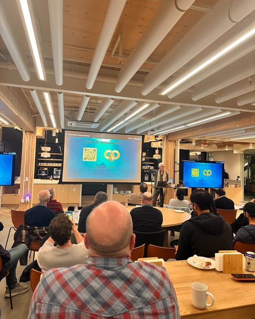 A photograph capturing an indoor presentation or meeting, showing a group of attendees seated around tables facing a large screen and a presenter standing to the side. The venue features a modern design with wood ceilings and distinctive white tubular lighting.