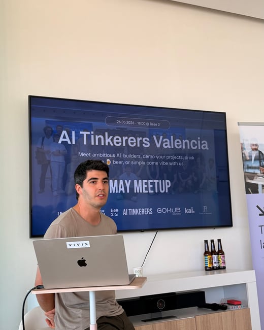 A man stands presenting next to a laptop in front of a large screen displaying an AI event slide.