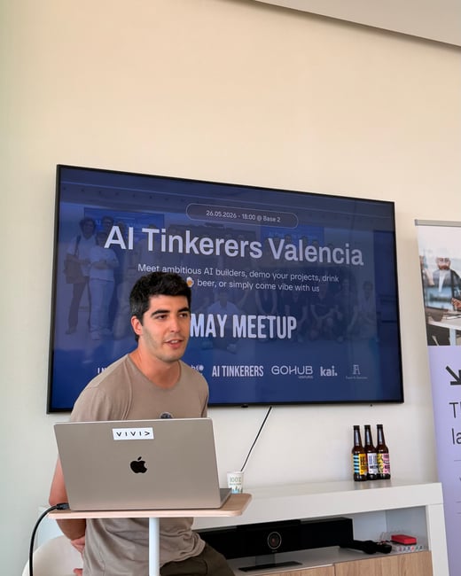 A man stands in front of a television screen displaying an 'AI Tinkerers Valencia' meetup presentation while using a laptop.