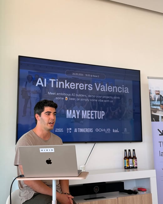 A man sits with a laptop in front of a large display screen presenting the 'AI Tinkerers Valencia' May meetup.