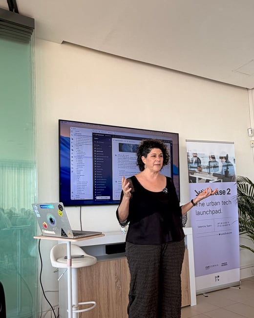 A woman stands giving a presentation in an indoor office setting, positioned next to a laptop stand and in front of a large monitor displaying software.