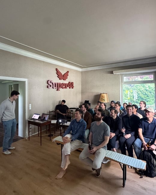 A group of people, primarily men, are seated on benches in a room, attending a presentation or meeting. A man stands to the left near a table with laptops, and a 'Super44' logo is mounted on the wall.