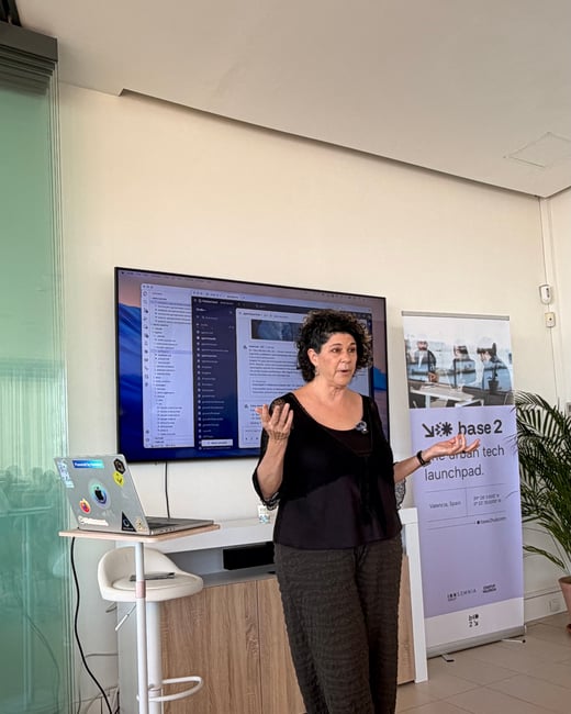 A woman stands in an office setting, presenting in front of a large screen displaying a software interface, with a laptop and promotional banner nearby.