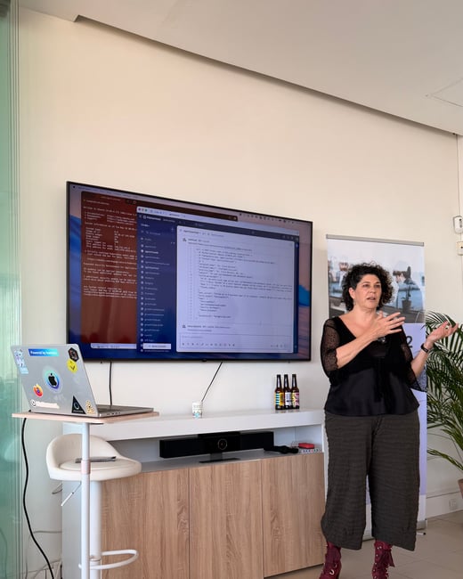 A woman stands gesturing during a presentation next to a large screen displaying terminal code and chat interfaces.