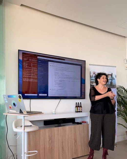 A woman stands and presents in front of a large monitor displaying terminal code and a chat interface.