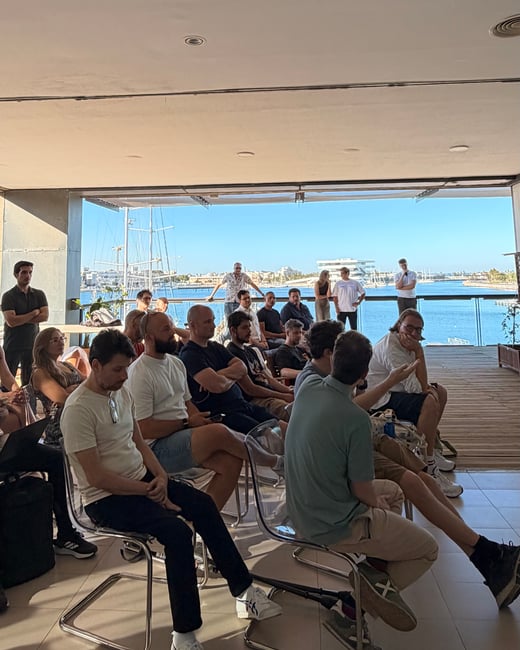 A group of people are seated indoors attending a presentation, with a large open window showing a marina filled with boats under a clear blue sky.