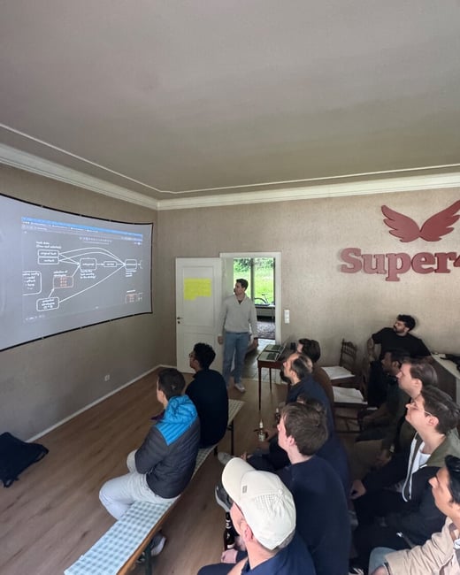 A group of people are gathered in a room for a presentation, watching a diagram projected onto a large screen while a man stands nearby.