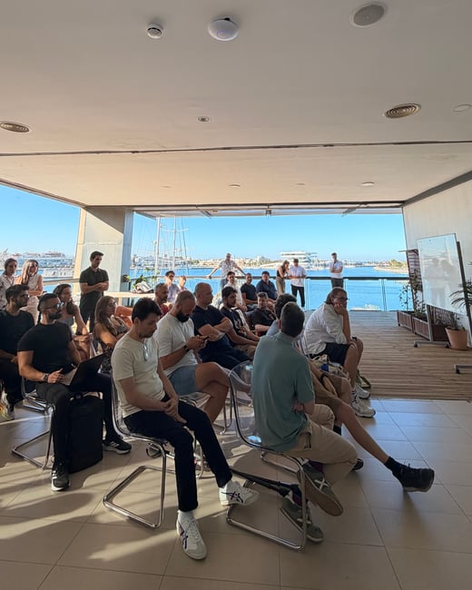 A group of people seated on a terrace attending a presentation with a scenic marina in the background.