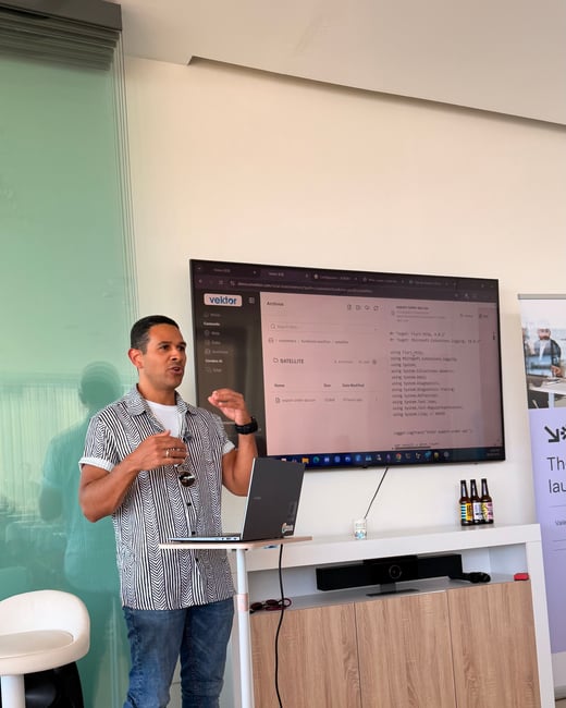 A man stands indoors giving a presentation next to a laptop stand, with a large screen displaying code behind him.