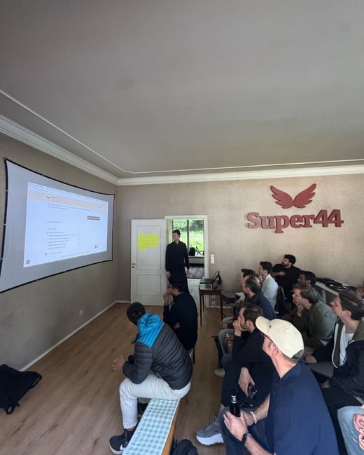 A group of people are gathered in a room, sitting and watching a presentation projected onto a screen. A prominent 'Super44' logo is mounted on the wall to the right.