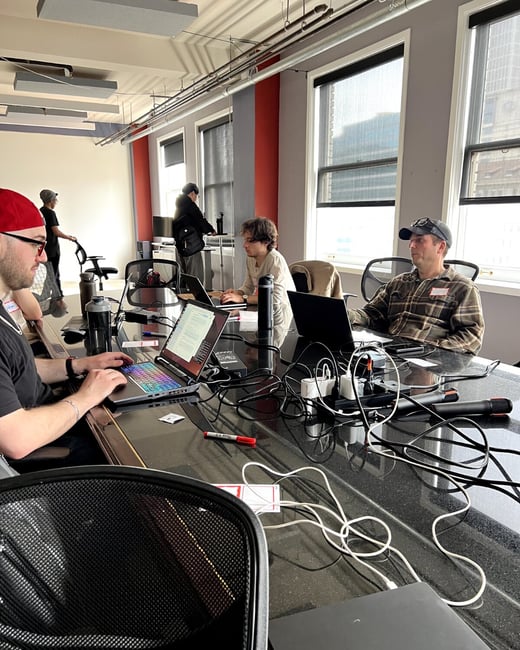 A group of people are working on laptops around a large black conference table in a modern office with large windows.
