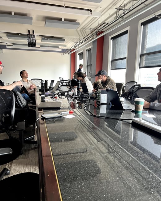 A group of people are gathered around a long conference table in an office setting, most of whom are working on laptops.