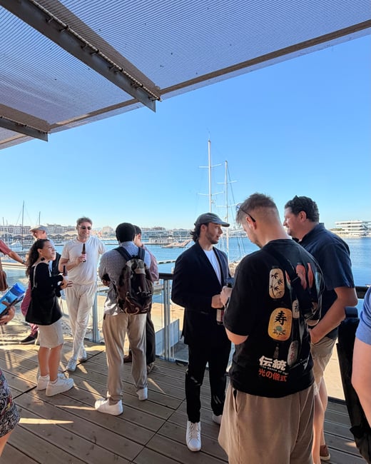 A group of people socializing on an outdoor wooden deck next to a harbor under a clear blue sky.