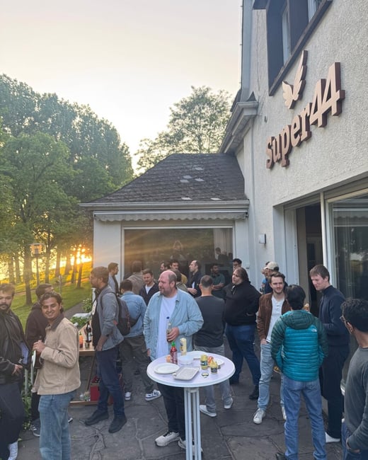 A group of people, primarily men, are gathered for a social event on an outdoor patio next to a building labeled 'Super44' during sunset.