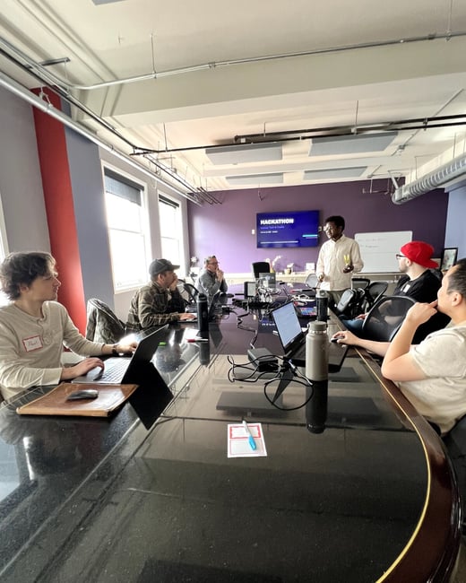 A group of people are gathered around a large, reflective conference table in a modern office, listening to a presentation about a hackathon.