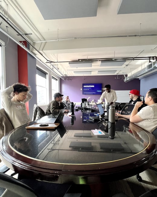 A group of people are gathered around a large, dark oval conference table in an office setting, with one person standing and presenting in front of a screen that reads 'HACKATHON'.