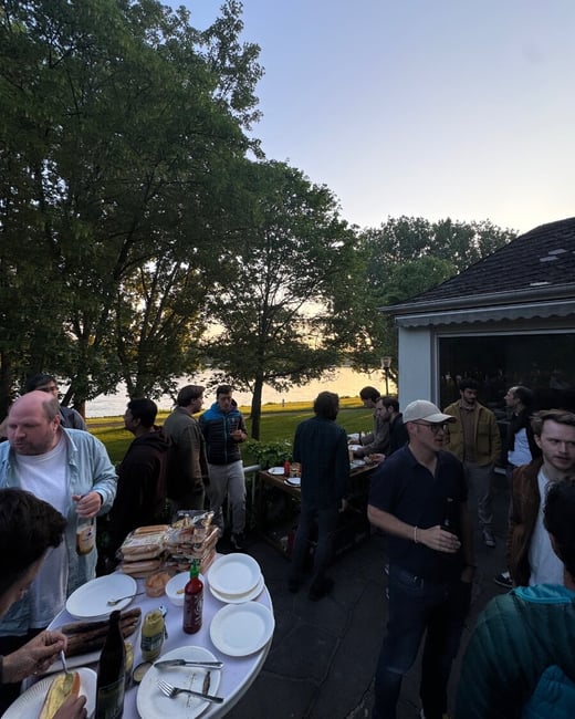 A group of people are gathered on an outdoor patio for a social event with food and drinks, overlooking a body of water at dusk.
