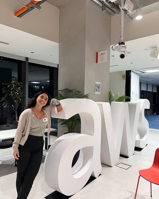 A woman smiles while leaning on large, white 3D letters spelling "aws" (Amazon Web Services) in a modern, well-lit corporate interior space.