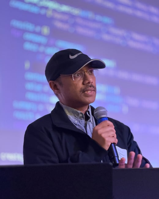 A man wearing a black Nike cap and glasses is speaking into a microphone during a presentation, with a screen showing blurred code in the background.