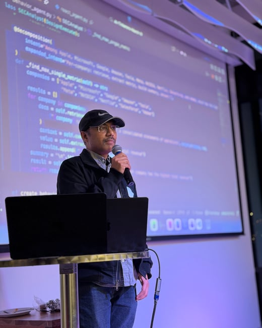 A man wearing a black Nike cap and glasses is giving a presentation with a microphone, standing in front of a screen displaying Python code.