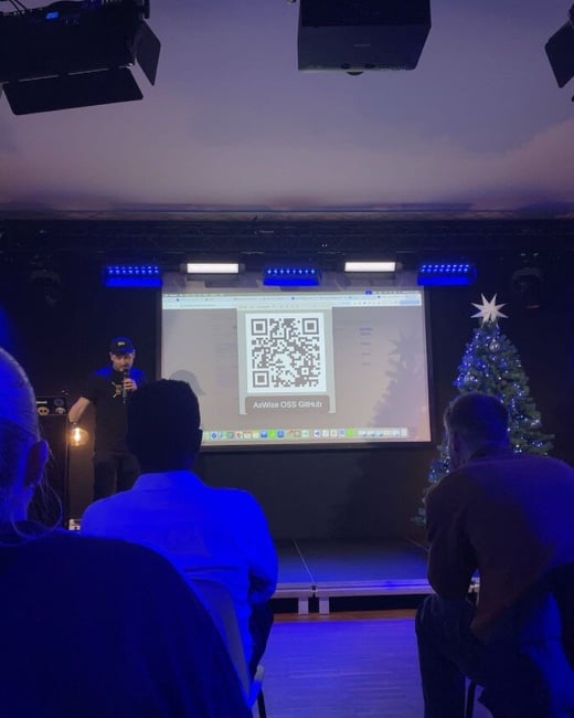 A photograph of a technical presentation taking place indoors, showing a speaker on a dimly lit stage next to a projected screen displaying a large QR code and the text "AxWise OSS GitHub," with several audience members visible in the foreground.