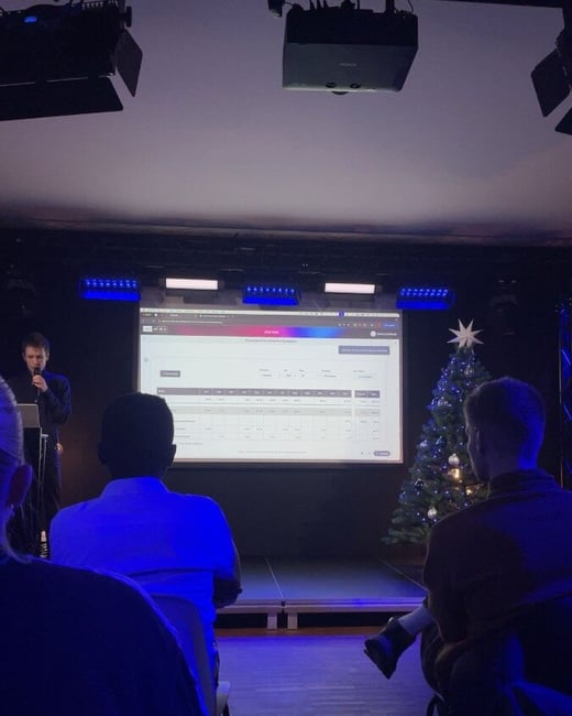 A photograph taken during an indoor presentation or event, showing a male speaker on a stage next to a large projection screen displaying a data dashboard, while several audience members watch from the foreground.