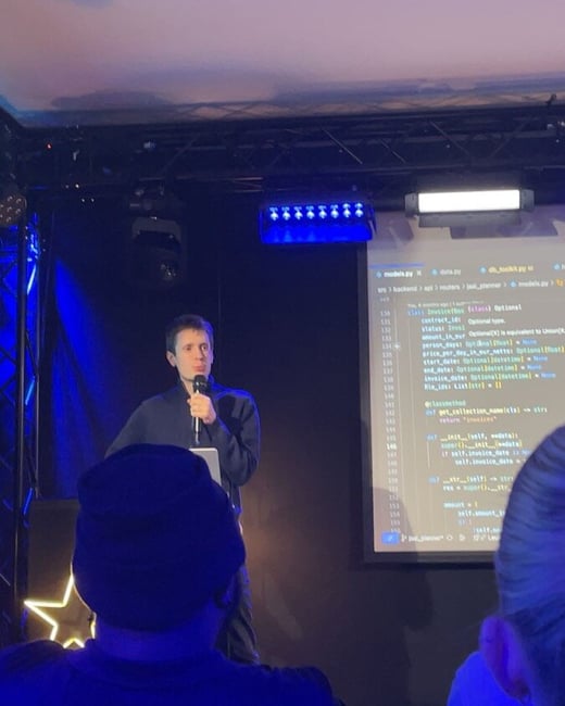 A low-light photograph captures a technical presentation where a man, illuminated by blue stage lights, speaks into a microphone while standing in front of a large screen displaying lines of Python code. The backs of audience members' heads are visible in the foreground.