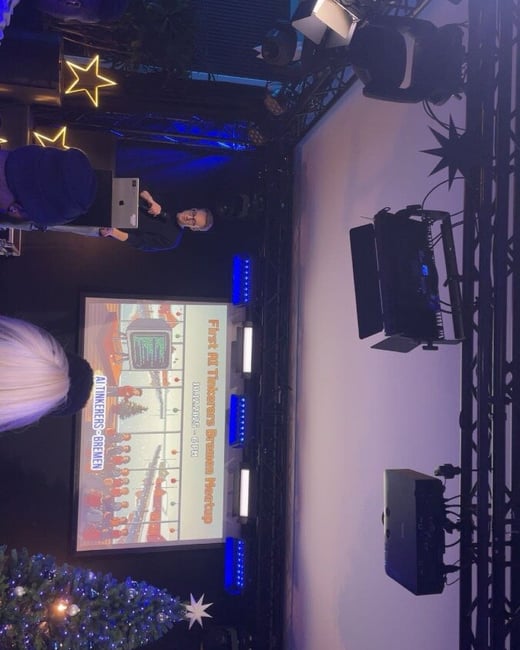 A low-light photograph captures an indoor presentation or meetup event, showing a male presenter standing next to a large projection screen displaying the title slide for the "First AI Tinkerers Bremen Meetup," with audience members visible in the foreground.