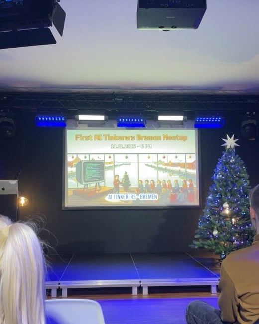 A photograph capturing an indoor event setup featuring a projection screen displaying a slide for the "AI Tinkerers Bremen Meetup," a decorated Christmas tree, and two attendees seated in the foreground facing the stage.