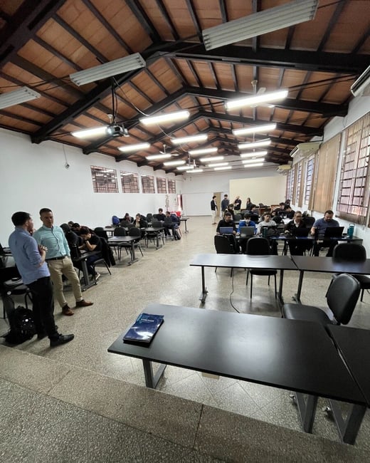 An indoor photograph of a large room with a vaulted wooden ceiling where several people are attending a workshop, some sitting at desks with laptops and others standing and talking.