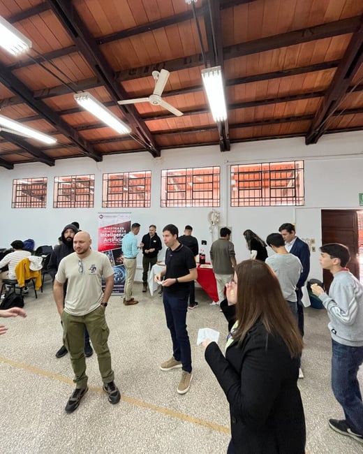 A group of people are gathered in a large room with high wooden ceilings for a social or networking event, with a banner for an Artificial Intelligence master's program visible in the background.