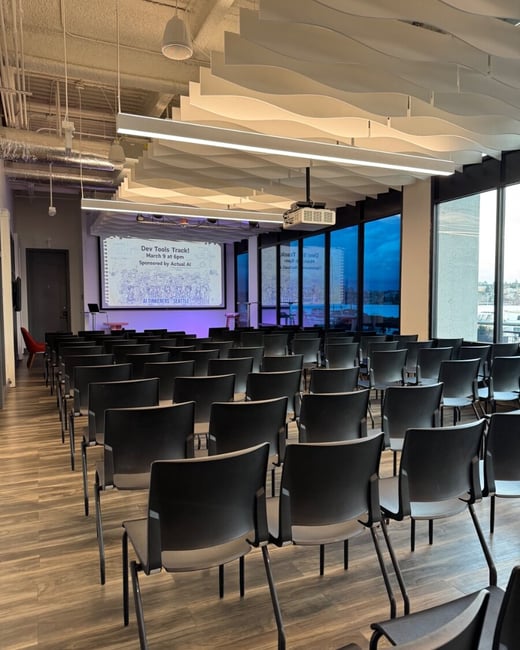 An empty presentation room with rows of black chairs facing a screen that announces a 'Dev Tools Track' event for AI Tinkerers Seattle.
