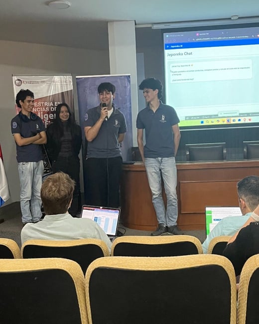 A group of students presenting a project called 'Jeporeka Chat' in an auditorium, with one student speaking into a microphone while others stand nearby.