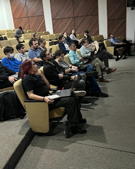 A group of people are seated in an auditorium, many with laptops open, appearing to be attending a presentation or lecture.