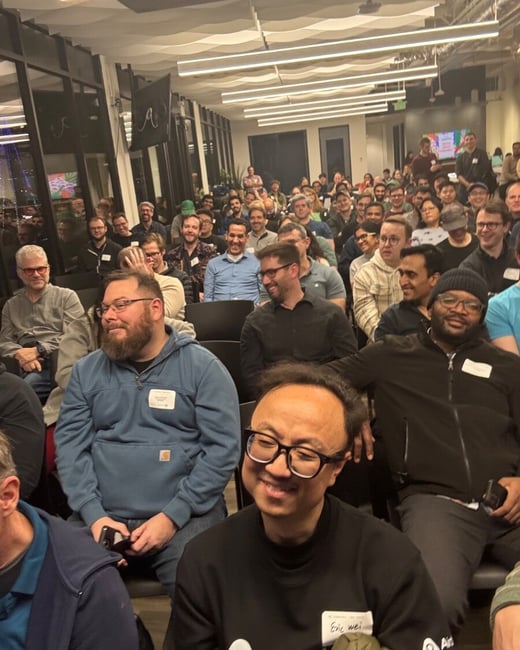 A vertical photo capturing a large audience seated indoors at an event or presentation, with many attendees smiling and looking toward the front of the room (where the camera is positioned). The setting appears to be a modern office or event space with distinctive wavy ceiling panels and large windows on the left.