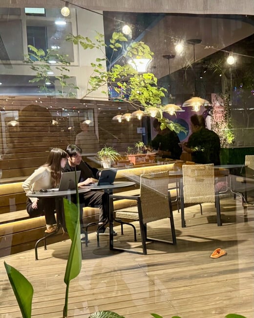 A candid photograph taken through a large glass window shows a young couple sitting at a table working side-by-side on laptops in an illuminated, semi-outdoor cafe seating area at night.