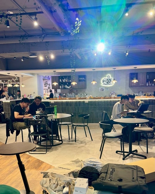 A candid photograph taken inside a modern cafe or co-working space, showing several people seated at tables working on laptops and devices, with a foreground table displaying merchandise including a black backpack and tote bags.