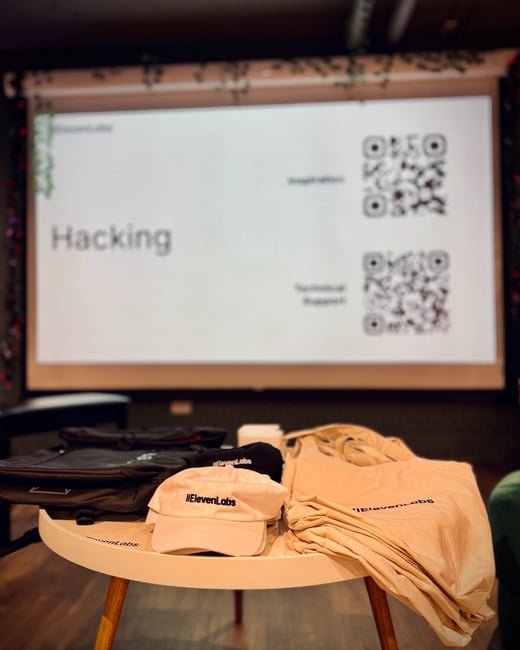 A low-light photo taken at an indoor event, featuring a table laden with ElevenLabs branded merchandise (hats, bags, and apparel) in the foreground, set against a large projection screen displaying a slide titled 'Hacking' and two QR codes in the background.