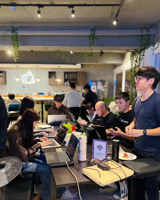 A group of people are gathered around a long table in a casual indoor setting, working intensely on multiple open laptops, suggesting a collaborative session or hackathon event.