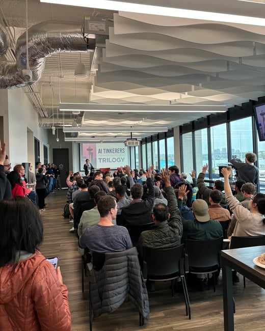 A photograph taken inside a modern event space shows a large audience attending a presentation, with many attendees raising their hands in response to the speaker or a prompt. The projection screen displays the title "AI TINKERERS TRILOGY" alongside an image of the Seattle Space Needle.