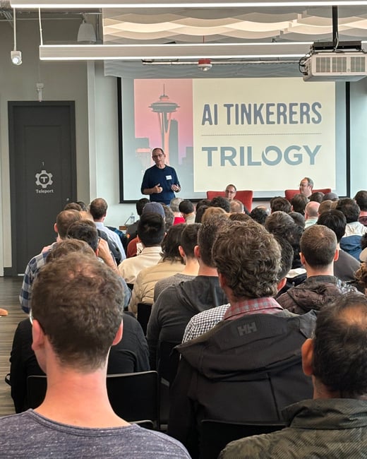 A photograph capturing an indoor event where a male speaker addresses a large, seated audience in front of a projection screen displaying the title 'AI TINKERERS TRILOGY' alongside a graphic of the Seattle Space Needle.