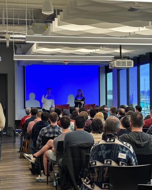 A photograph capturing an indoor event or presentation where a large audience is seated, facing two speakers standing in front of a massive, bright blue projection screen. The venue features modern architecture, including wavy acoustic ceiling panels and large windows offering a view of a waterfront.