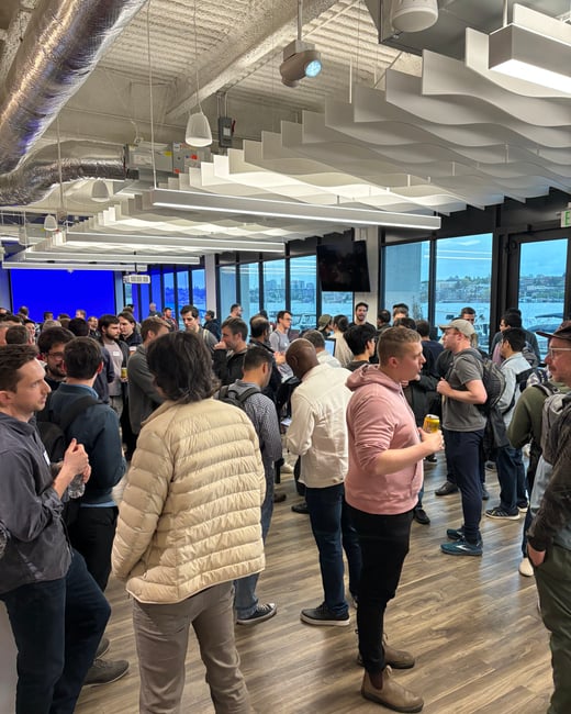 A photograph capturing a large group of people mingling and networking at a crowded indoor event space, featuring modern architecture, high ceilings, and floor-to-ceiling windows overlooking a waterfront cityscape.